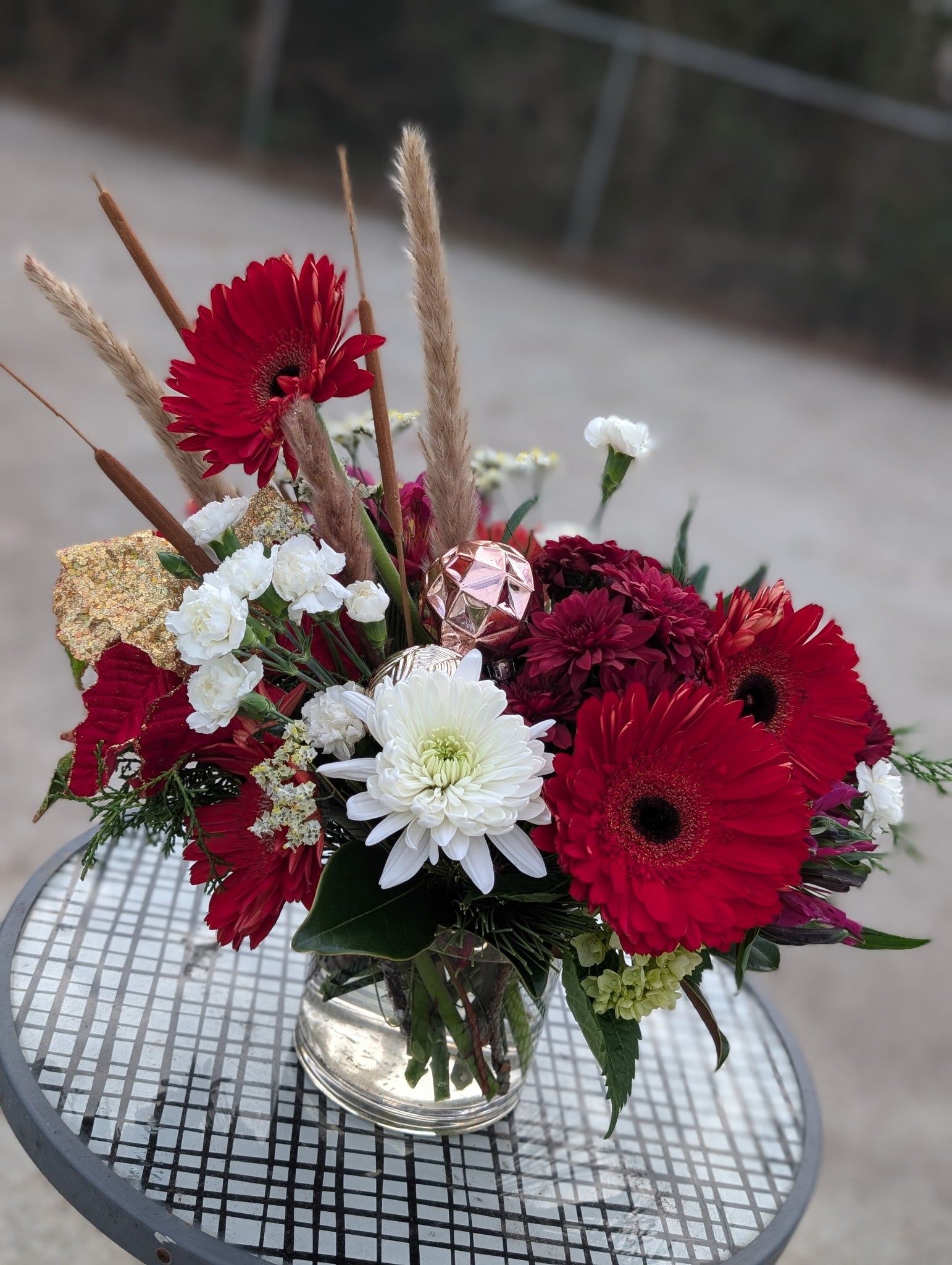 Holiday arrangement with red gerbera daisies, red carnations, red mums, white cushion mum, white mini carnations, pampas grass, cattail stems, gold-dusted hydrangea, and greenery in a clear glass vase.