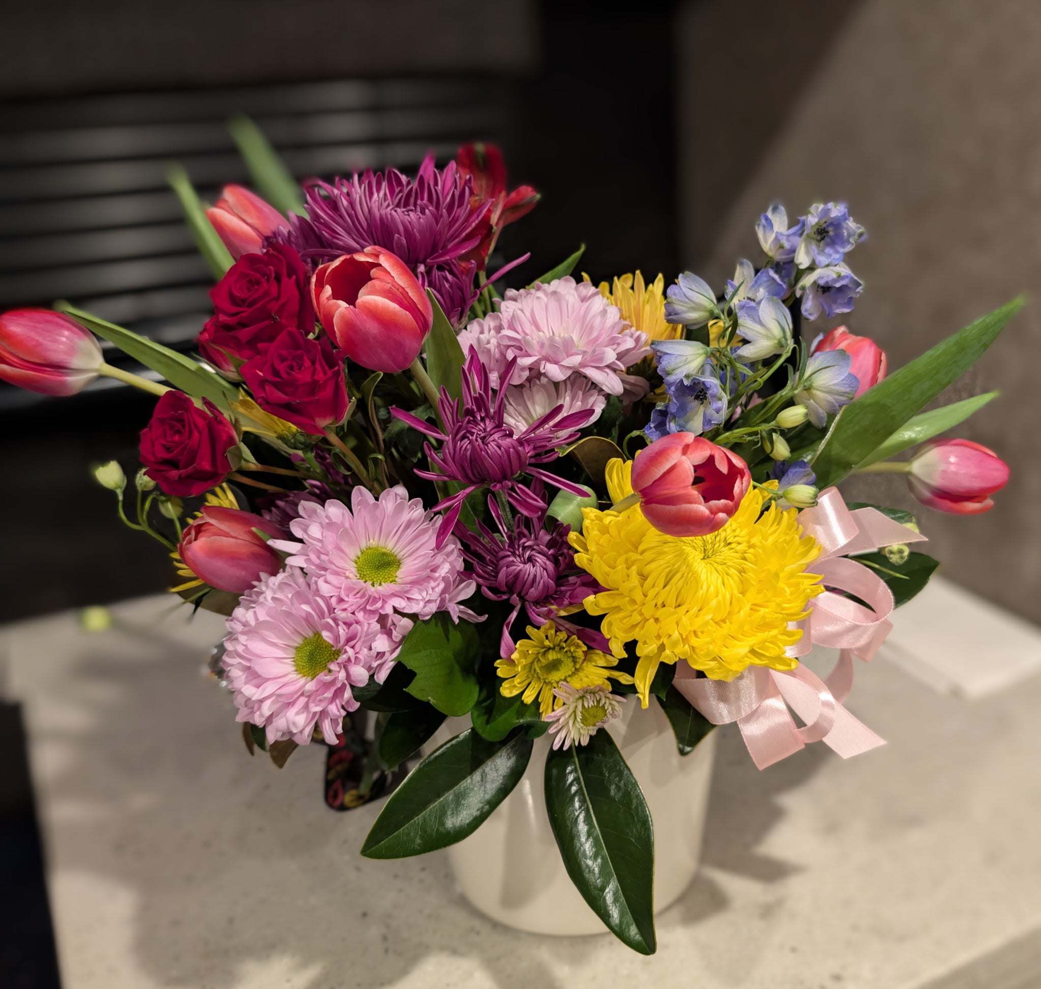 Colorful mixed flower arrangement with tulips, roses, chrysanthemums, delphinium, and greenery in a white ceramic vase, finished with a soft pink ribbon.