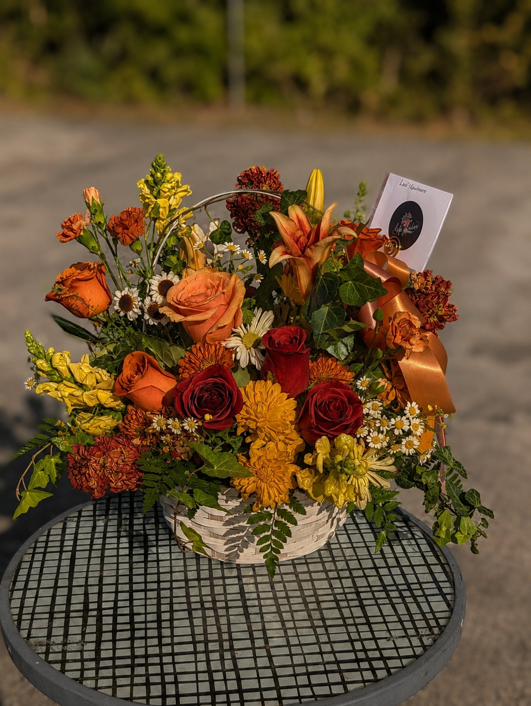 A handcrafted fall flower basket featuring orange and red roses, lilies, chrysanthemums, snapdragons, daisies, seasonal blooms, and lush greenery in a white woven basket, perfect for autumn gifting.