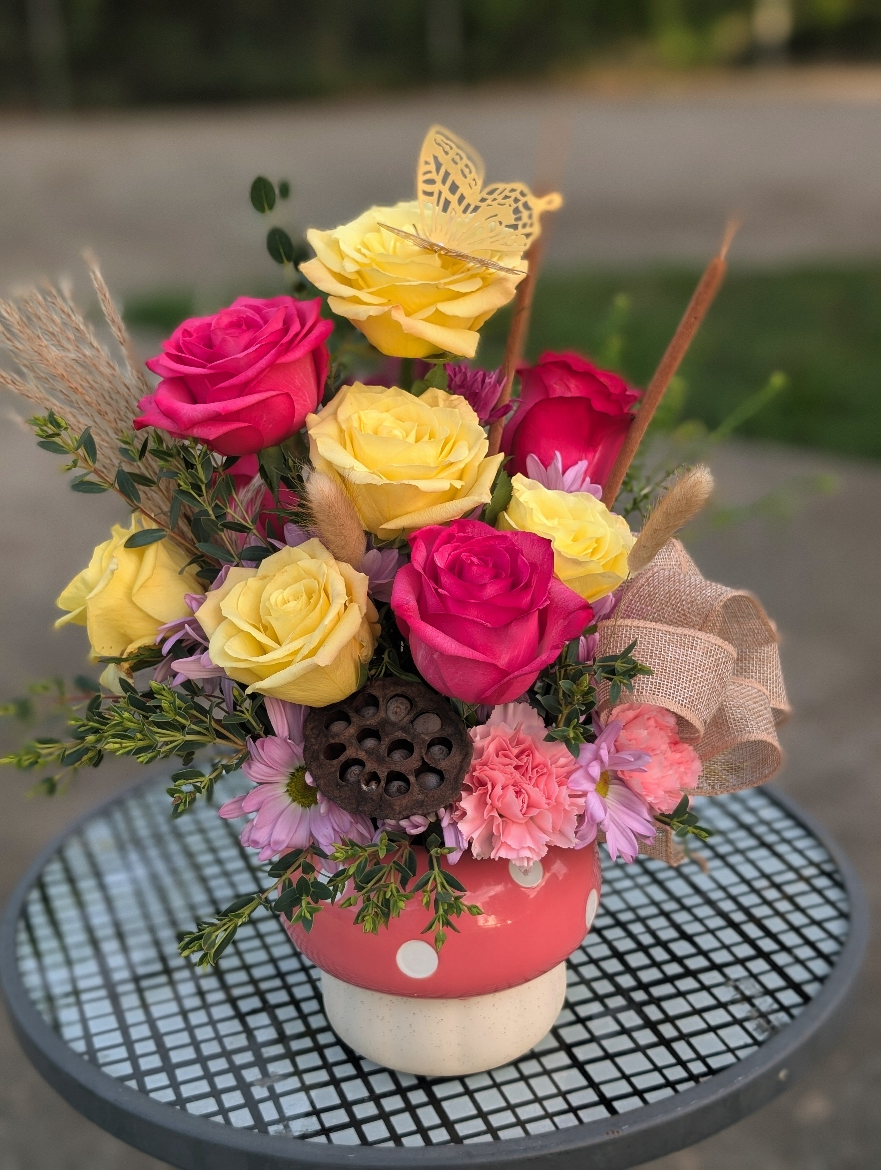 Garden-style flower arrangement featuring yellow roses, pink roses, carnations, daisies, and a decorative butterfly in a pink mushroom keepsake vase.