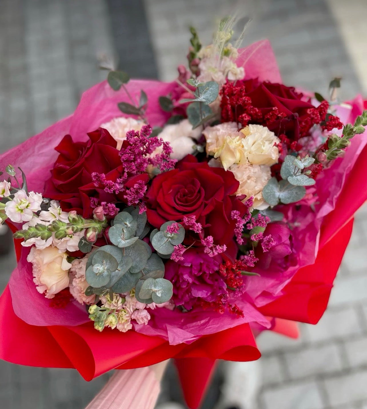 Hand-tied bouquet of red roses and soft pink blooms wrapped in pink and red paper, accented with eucalyptus and seasonal filler flowers for a romantic, gift-ready look.