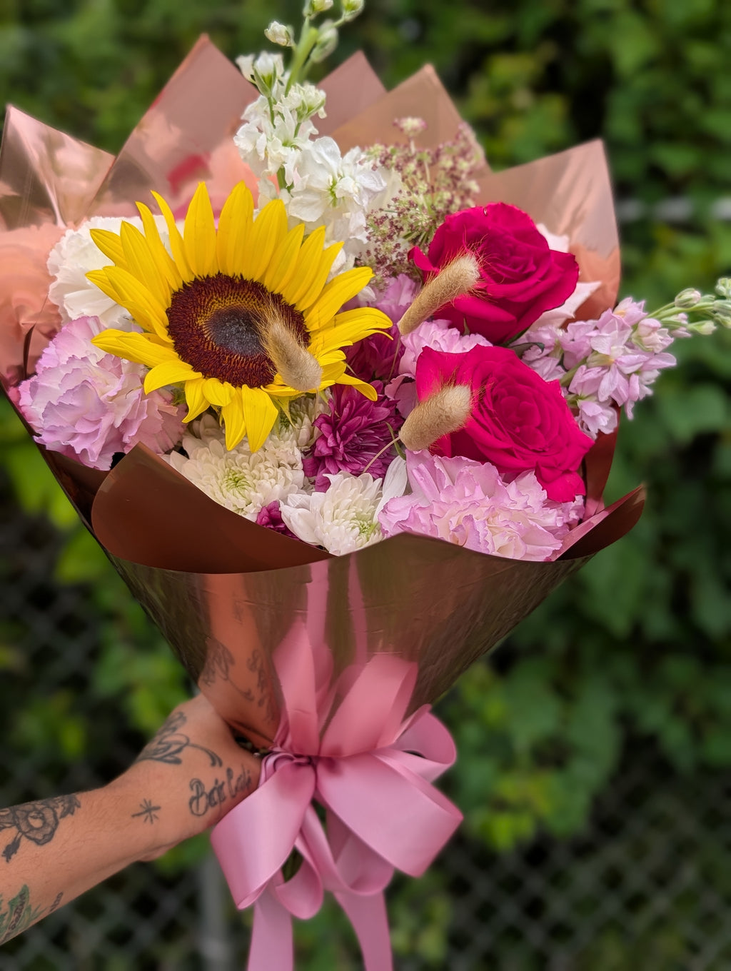 Hand-tied bouquet featuring a sunflower, hot pink roses, pink stock, carnations, and seasonal blooms wrapped in rose gold paper with a pink satin ribbon.