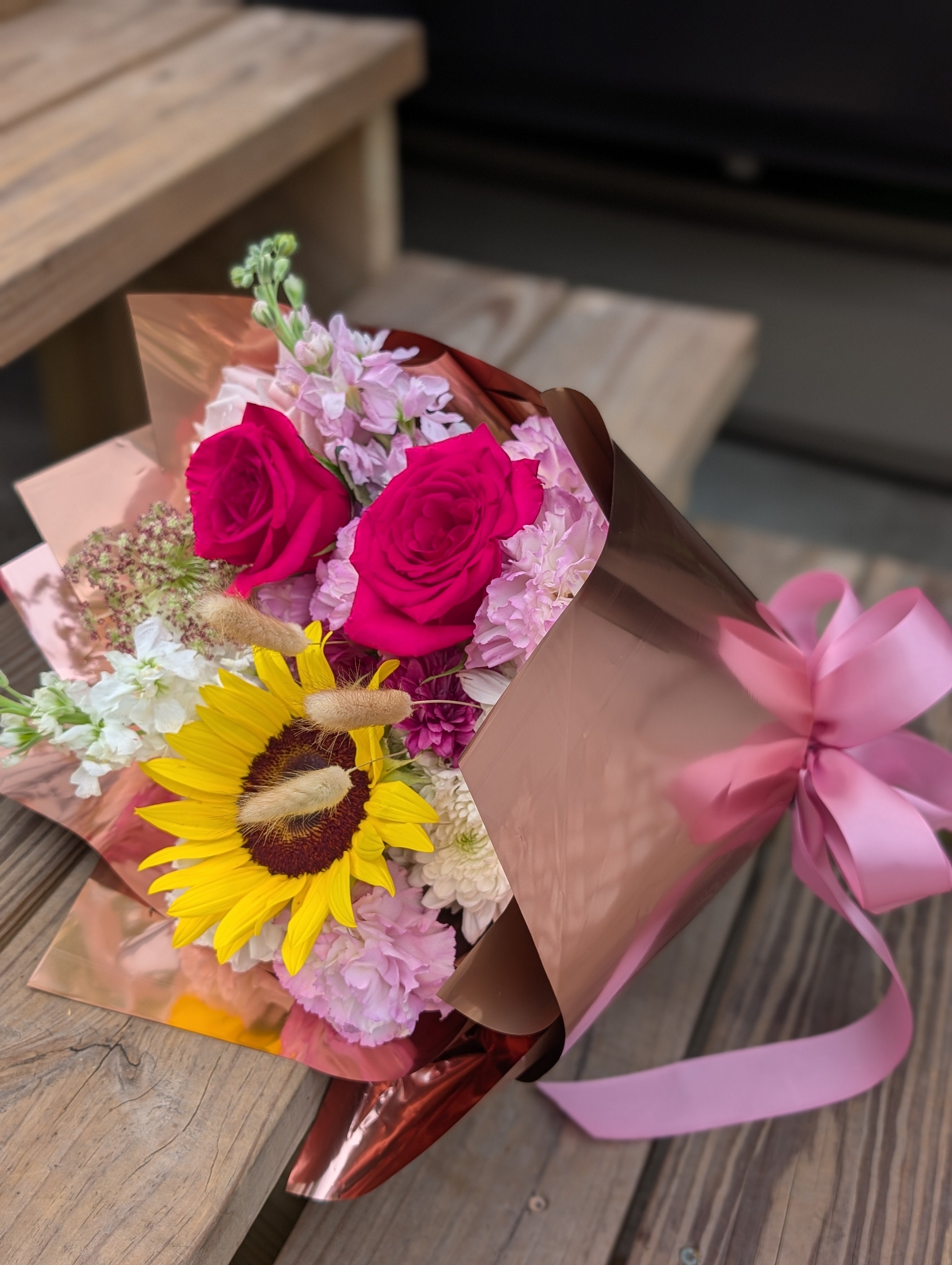 Hand-tied bouquet with sunflower, hot pink roses, pink stock, carnations, and seasonal flowers wrapped in rose gold paper with a pink ribbon.